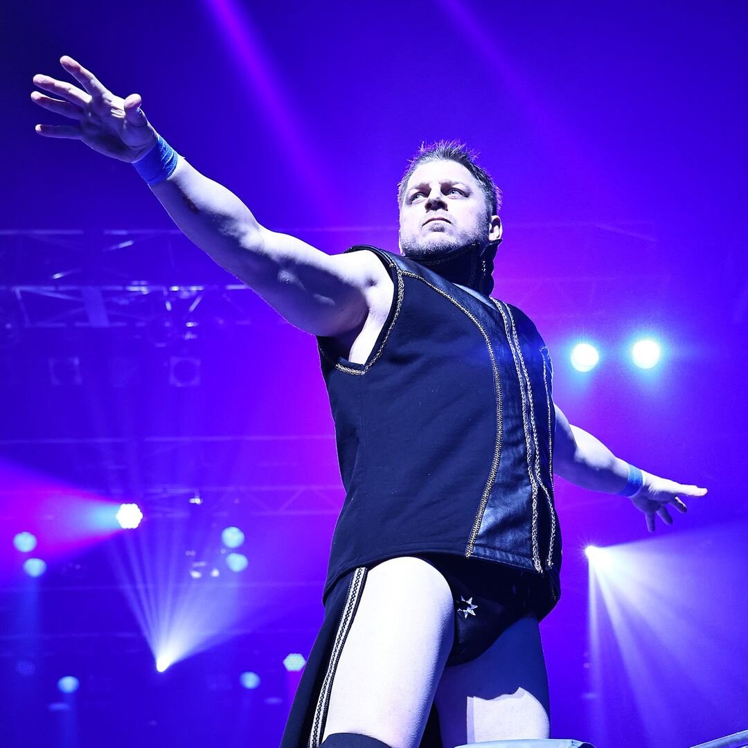 Rionne Fujiwara raising his arms in the ring under purple lighting